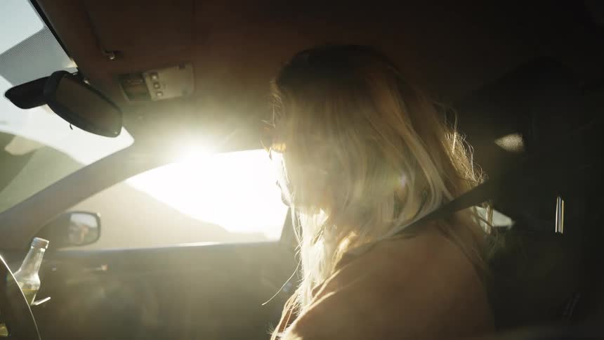 A blonde woman wearing sunglasses drives her car as the golden hour sun shines brightly, creating a cinematic lens flare