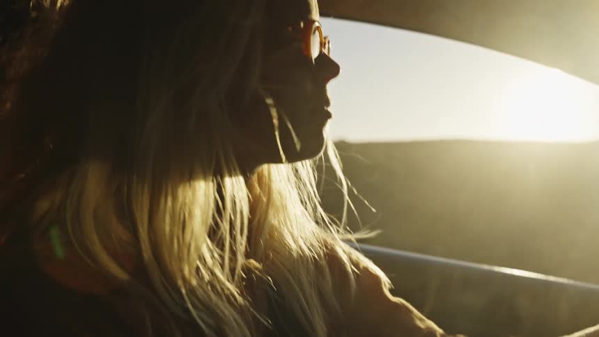 A beautiful blonde woman with sunglasses drives her car during a stunning golden hour sunset as her hair blows in the wind