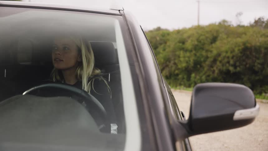 A young blonde woman gets into her car, fastens her seatbelt, and displays a moment of frustration before preparing to drive