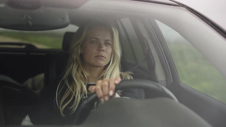 A focused blonde woman sits thoughtfully behind the wheel of her car as she drives down the road