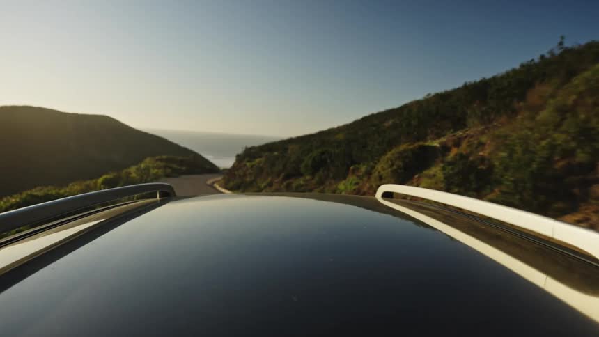 A scenic point-of-view from a car roof driving along a winding coastal highway through rolling hills at golden hour