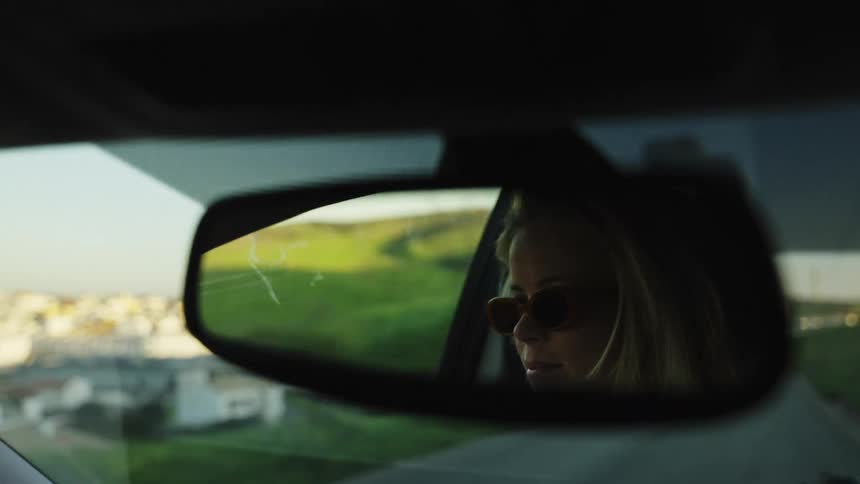 A stylish blonde woman wearing sunglasses is reflected in the rearview mirror as she drives through a scenic landscape