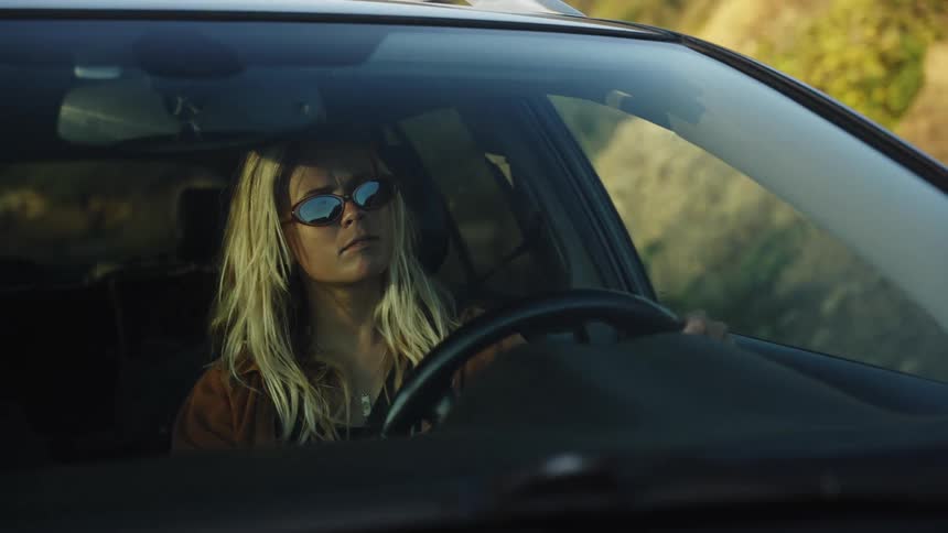 A blonde woman in sunglasses drives a car through a sunny landscape, seen from the front windshield
