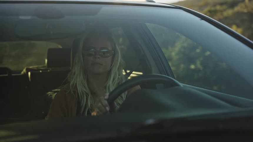 A blonde woman in sunglasses drives her car down a scenic road during a beautiful golden hour sunset