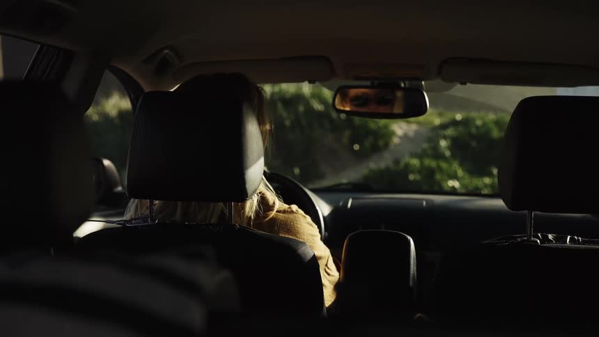 A woman gets into the driver's seat and puts on her seatbelt, filmed from the back seat of the car at sunset