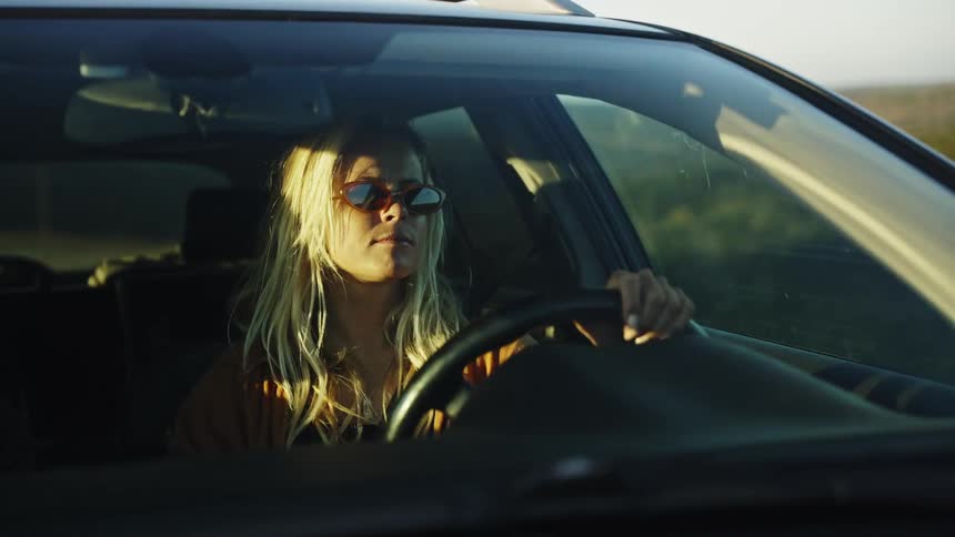 A stylish blonde woman wearing sunglasses drives her car through the countryside during a beautiful golden hour