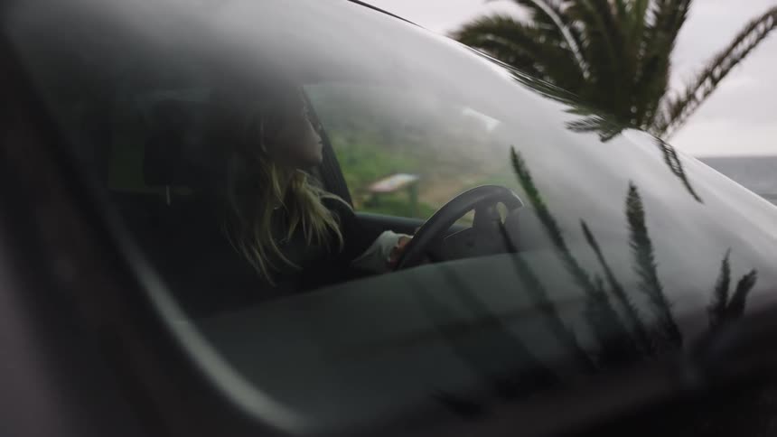 A blonde woman gets into the driver's seat of her car parked by the coast on an overcast day