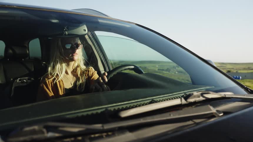 A stylish young blonde woman in sunglasses drives her car on a scenic road trip through a sunny, green countryside