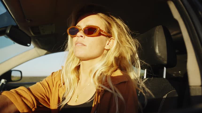 A stylish blonde woman in sunglasses gets out of her car to enjoy the sunny weather during a scenic road trip