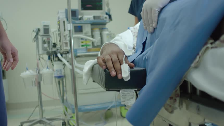 In a sterile operating room, a patient's hand rests on a gurney with a pulse oximeter attached as medical personnel attend to them