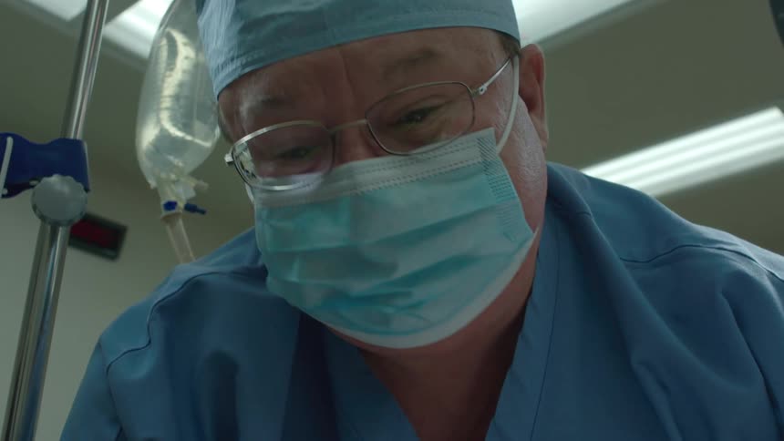 A surgeon in a mask and scrubs looks down at an unconscious patient on life support in the operating room