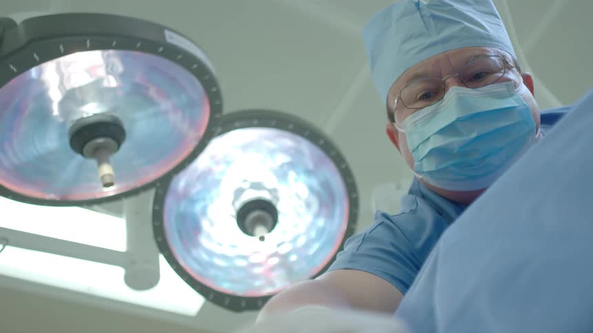 A patient's point of view shows a surgeon in a mask and gloves leaning over them in a brightly lit operating room