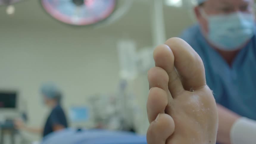 A medical team prepares a patient for surgery, with the camera slowly shifting focus from a nurse attaching a monitor to the surgeon holding the patient's foot
