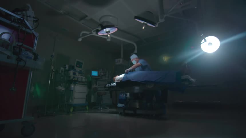 A blurry, low-angle, wide shot of a surgeon operating on a patient in a dark and dramatic operating room