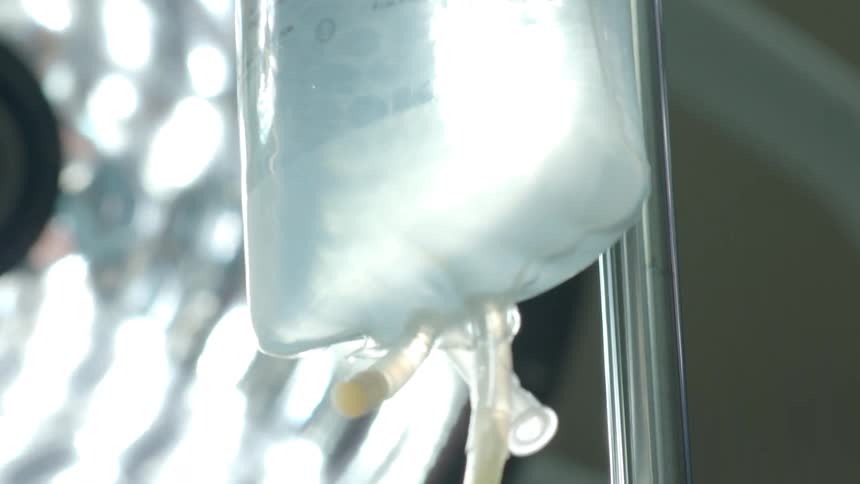 A slow pan down reveals a close-up of an IV drip bag and chamber administering fluids in a hospital operating room
