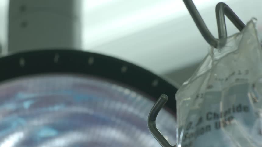 An IV bag of sodium chloride hangs on a stand with a bright surgical light in the background of an operating room