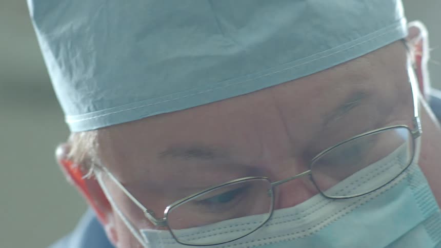 A surgeon in a mask and glasses focuses intently on a procedure under a bright operating room light