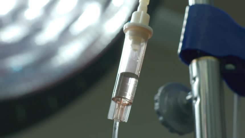 A close-up view of an intravenous drip in a hospital operating room, with a bright surgical light out of focus in the background