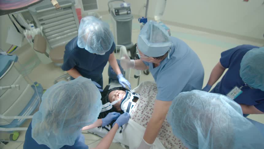 From a high-angle perspective, a team of doctors and nurses in an operating room work together on a patient with a neck brace and breathing tube