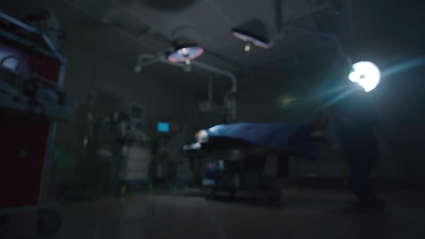 An out-of-focus wide shot of a doctor with a flashlight slowly approaching a patient on an operating table in a dark hospital room