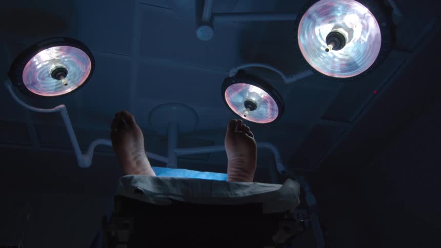 A low-angle view shows the feet of a deceased person on a gurney under bright surgical lights in a morgue