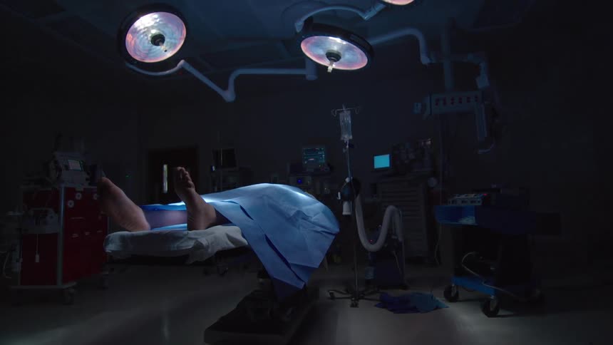 Slow dolly out reveals a patient on a table in a dark and empty operating room lit by surgical lights