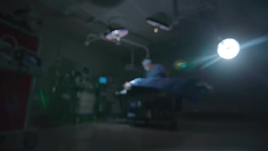 An out-of-focus, dramatic wide shot of a surgeon performing a procedure on a patient in a dark operating room