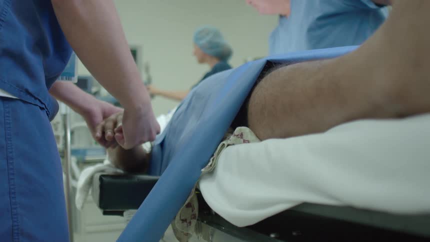 A nurse gently holds a patient's hand in a busy operating room, with a slow camera move to the patient's foot
