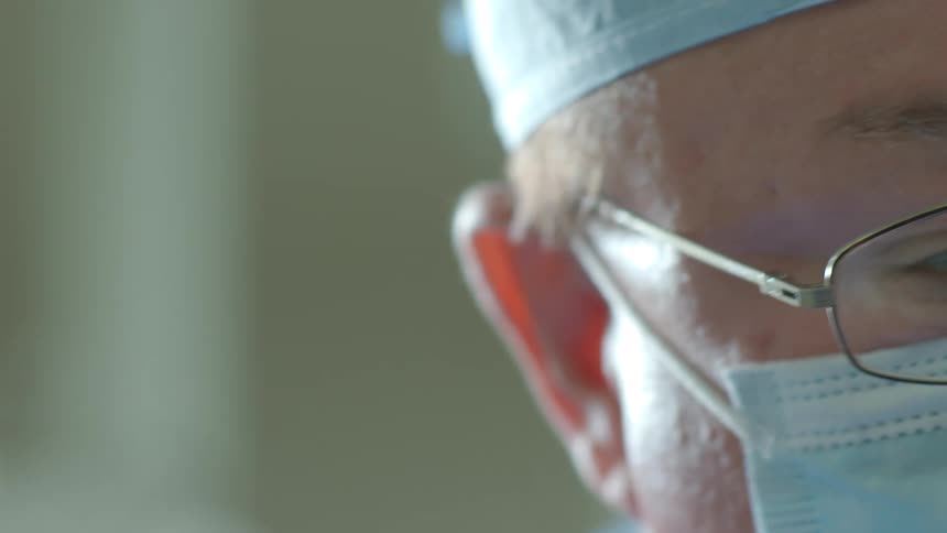 A slow pan reveals the concentrated face of a surgeon wearing a mask and glasses in an operating room