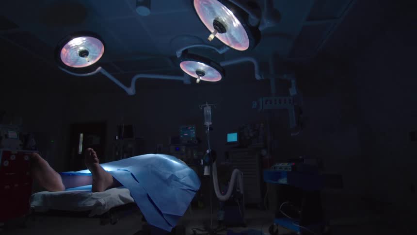 A slow pull-back shot reveals a patient lying on a gurney in a dark and sterile operating room under bright surgical lights