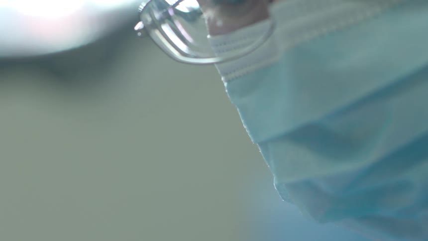 An intense slow motion close-up of a surgeon's face wearing a mask and glasses while performing a procedure in the operating room