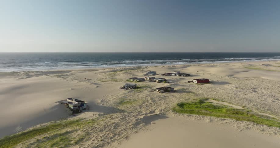 A high-angle static shot reveals a small, isolated settlement of weathered shacks amidst expansive sand dunes along a deserted coastline