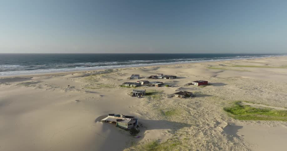 An aerial view shows a small, isolated settlement of beach shacks nestled among vast sand dunes next to the ocean on a clear day