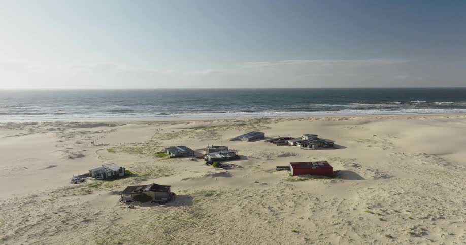 A static aerial view shows a small, isolated village of shacks on a vast sandy beach next to the ocean