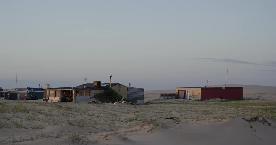 A quiet cluster of rustic, off-grid shacks sits among the sandy dunes at dusk