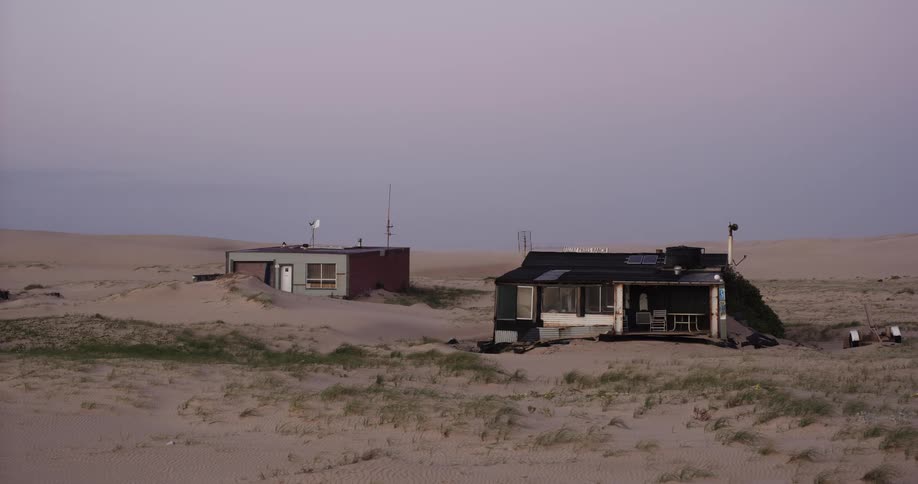 Weathered and abandoned houses are slowly being swallowed by a vast, desolate sand dune landscape at twilight