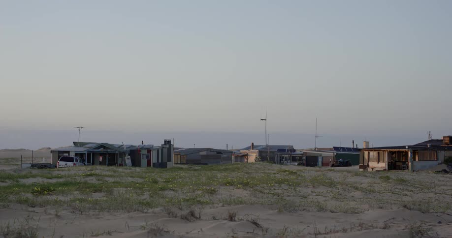 A still shot of a rustic, off-the-grid village with simple shacks on a sandy landscape at dusk