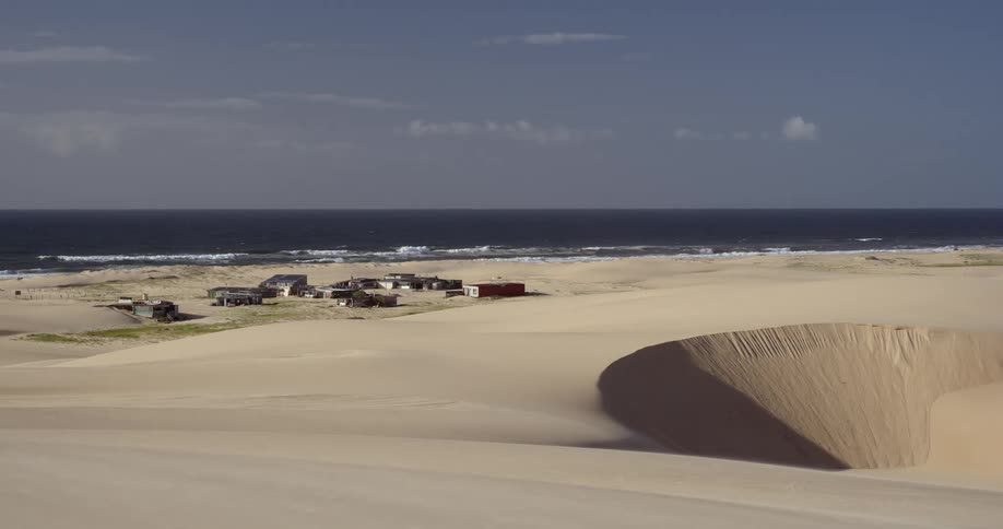 An isolated village is nestled between vast sand dunes and the ocean on a remote coastline