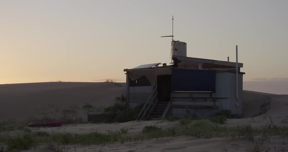 A rustic, off-grid shack made of corrugated iron sits peacefully among vast sand dunes at sunset