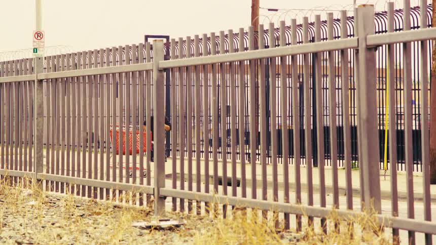 A man pushes a red shopping cart down an urban sidewalk, viewed from behind a tall metal security fence