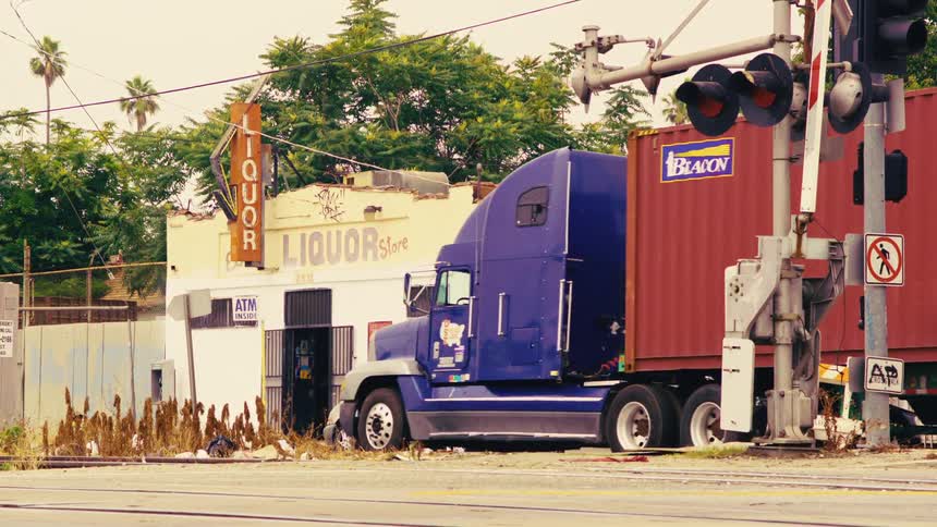 A run-down liquor store sits next to a railroad crossing as traffic and pedestrians pass by on a city street