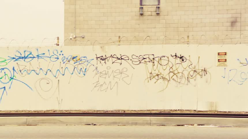A smooth panning shot from a moving vehicle captures a long graffiti-covered wall alongside a busy urban street