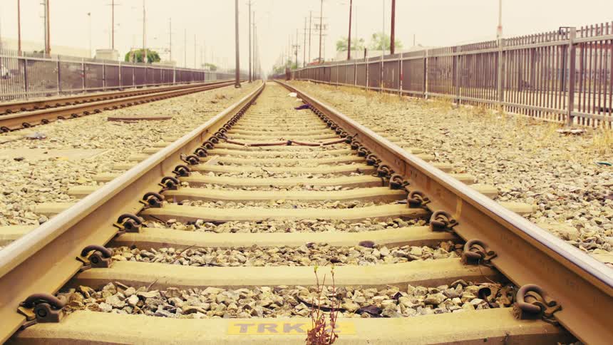 Empty railroad tracks stretch towards a vanishing point under a hazy, warm sky in a desolate industrial landscape