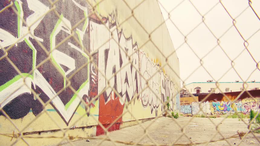 A static shot of a graffiti-covered wall in a desolate urban lot, viewed through a chain-link fence