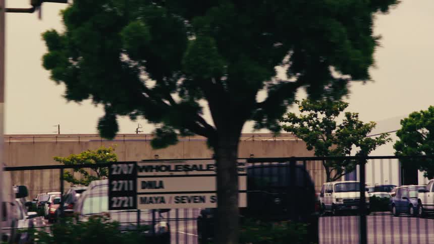 A smooth tracking shot from a moving vehicle captures a long building wall, landscaping, and a parking lot in an urban industrial area