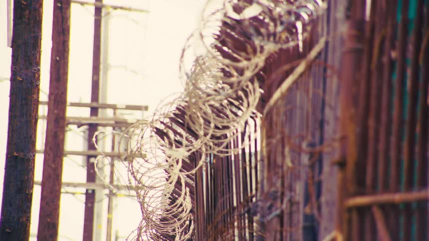 A close-up rack focus shot reveals sharp, coiled razor wire and barbed wire on a rusty security fence