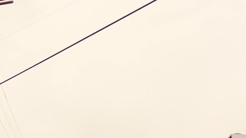 A low-angle drive-by shot looking up past power lines and traffic signals to a 'Compton' street sign against an overcast sky