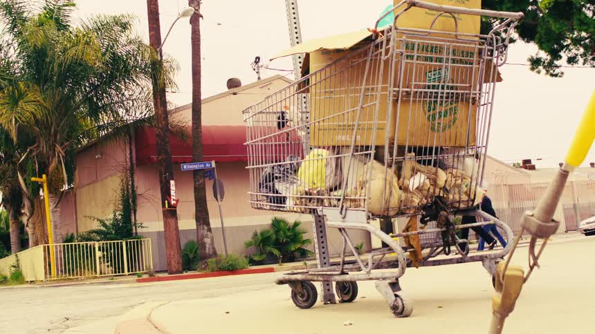 A homeless person's shopping cart filled with belongings sits on a city street corner as people and cars pass by in the background