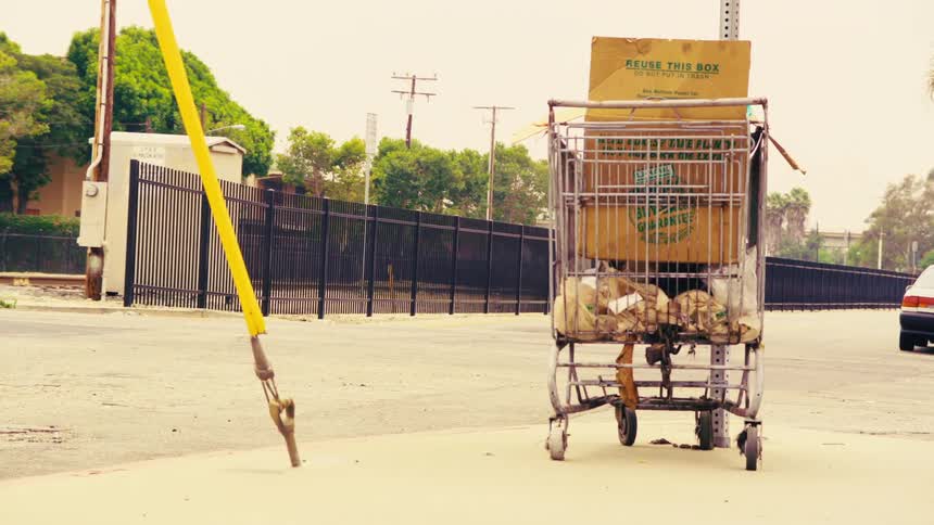 An abandoned shopping cart filled with cardboard boxes and personal belongings sits on a sidewalk in a desolate urban area