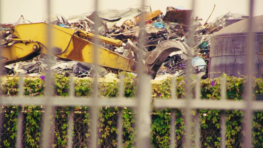 A large yellow mechanical claw sorts through a massive pile of scrap metal in slow motion at a recycling facility